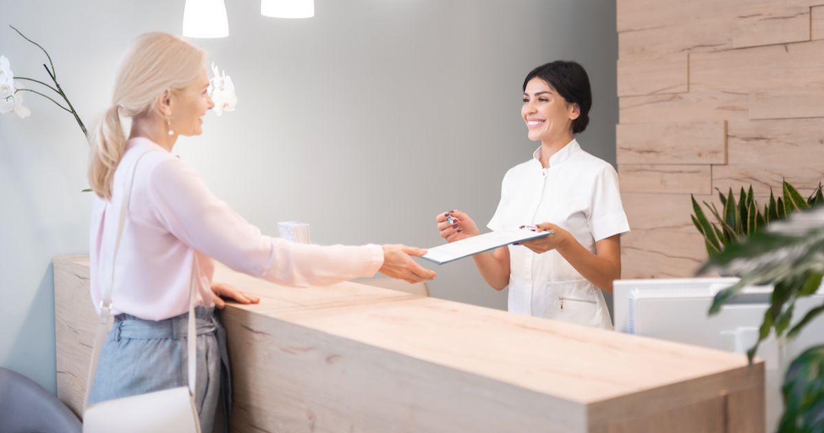 Die Rezeptionistin einer Zahnarztpraxis übergibt einer Patientin ein Formular.