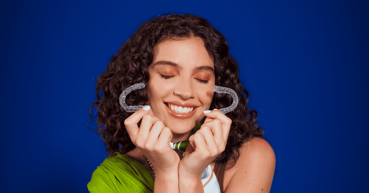 Chica sonríe a la cámara con los ojos cerrados mientras sujeta un alineador Invisalign® con la mano.