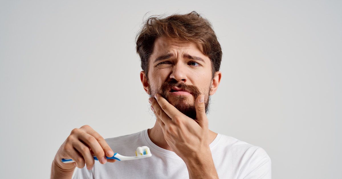 Chico se toca la mandíbula como signo de dolor y sujeta un cepillo de dientes con pasta sobre fondo blanco.