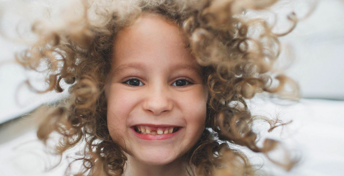 ein kleines Mädchen mit Locken lacht in die Kamera und zeigt ihre Milchzähne und Zahnlücke.