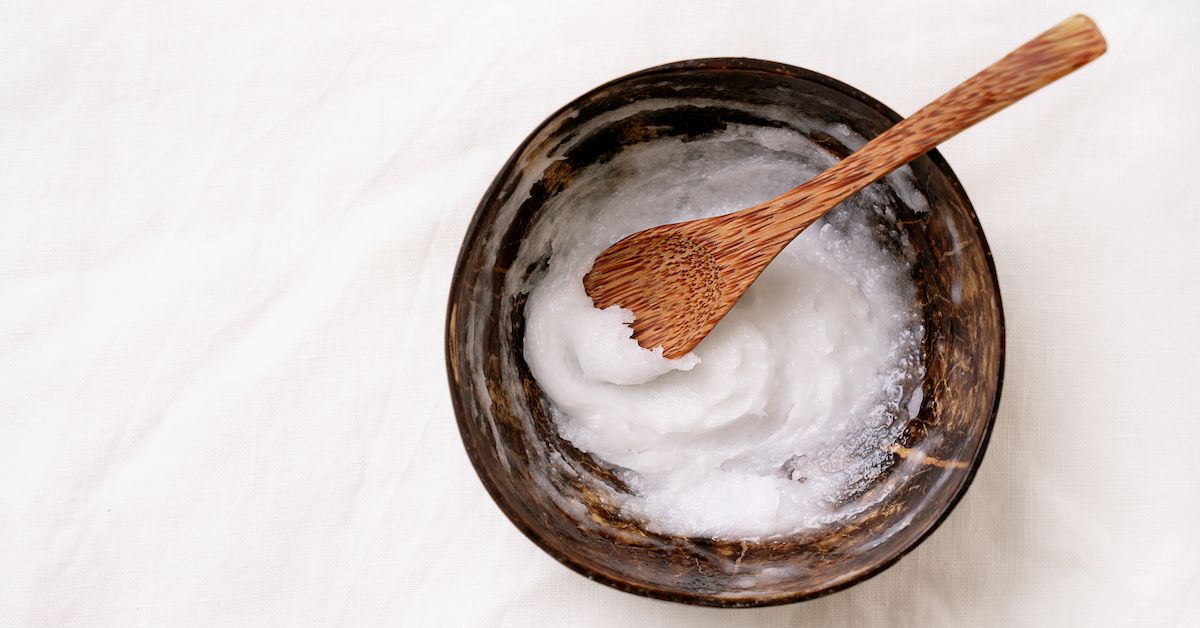 Un tarro y una cuchara de madera de coco que contiene aceite de coco dentro sobre fondo blanco.