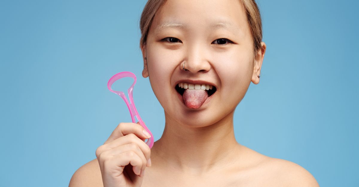 Una chica sujeta un raspador de lengua de plástico rosa con la mano y saca la lengua sobre fondo azul.