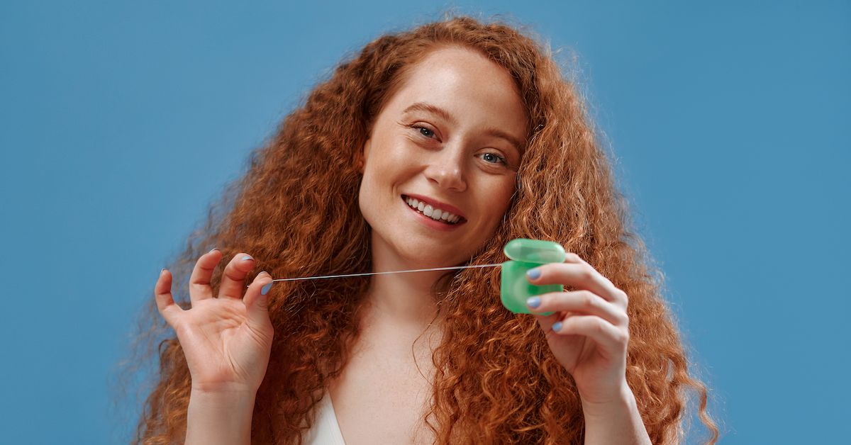 Una chica peliroja que sonríe y muestra hilo dental con sus manos sobre fondo azul.