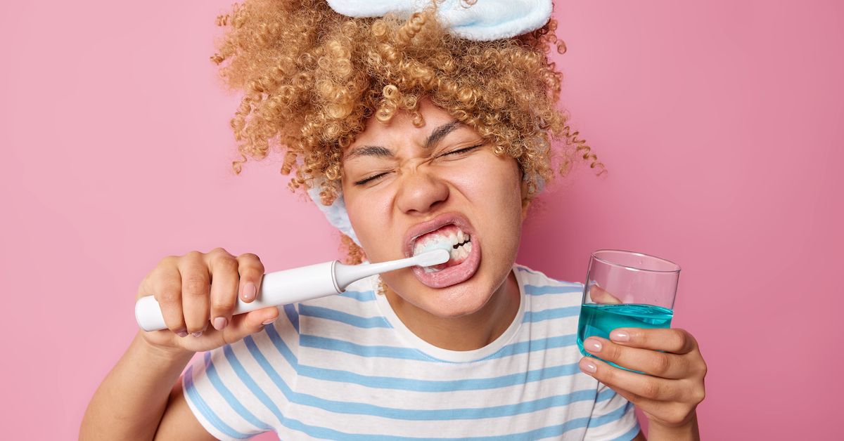Una chica que se lava los dientes con un cepillo eléctrico y con la otra mano sujeta un vaso azul de enjuague bucal.